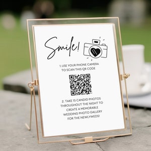 May include: A white sign with black text and a black and white camera icon with a heart inside. The sign says "Smile!" and has instructions to use a phone camera to scan a QR code to take candid photos at a wedding.