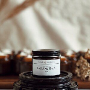 May include: A small brown glass jar with a black lid and a white label that reads "sunnah apothecary" and "UNSCENTED TALLOW BALM 1.5 OZ". The jar is sitting on a dark brown wooden pedestal.