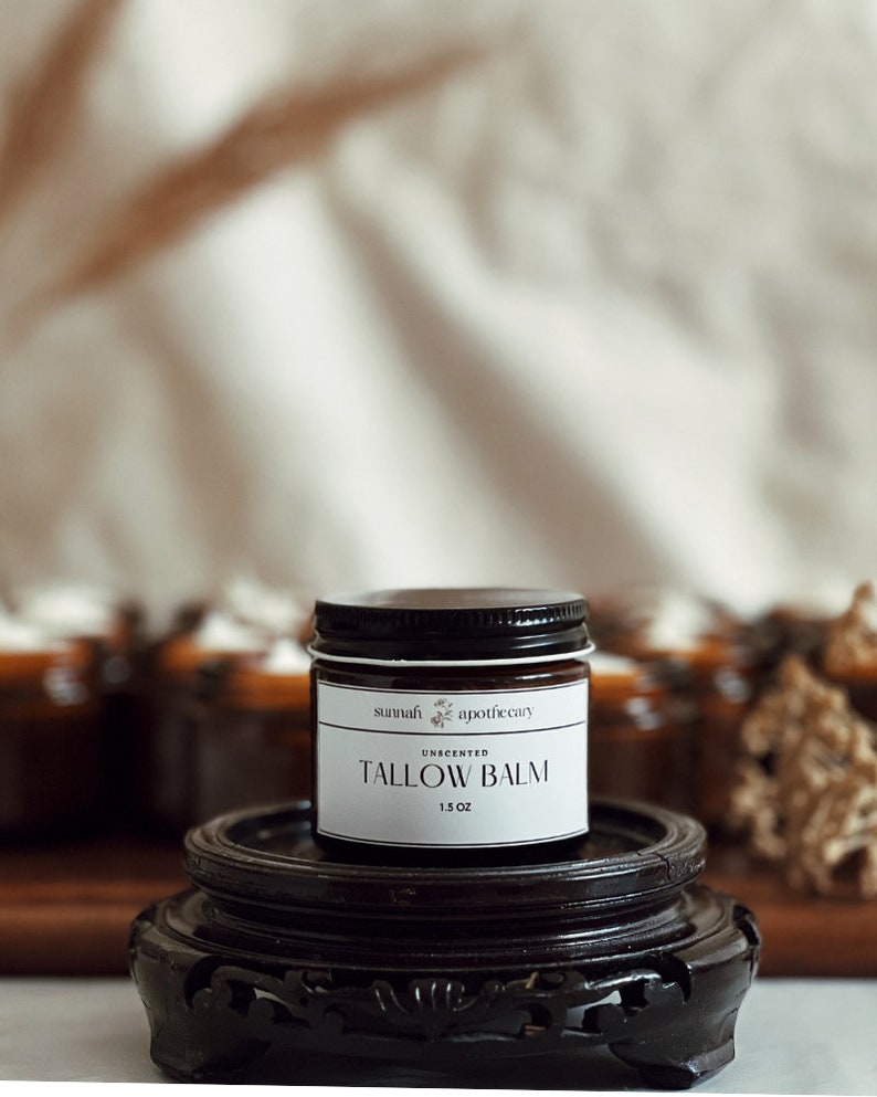 May include: A small brown glass jar with a black lid and a white label that reads "sunnah apothecary" and "UNSCENTED TALLOW BALM 1.5 OZ". The jar is sitting on a dark brown wooden pedestal.