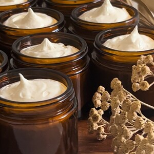 May include: Multiple amber glass jars filled with a white cream product. The jars are arranged in a row on a wooden surface with dried flowers in the background.