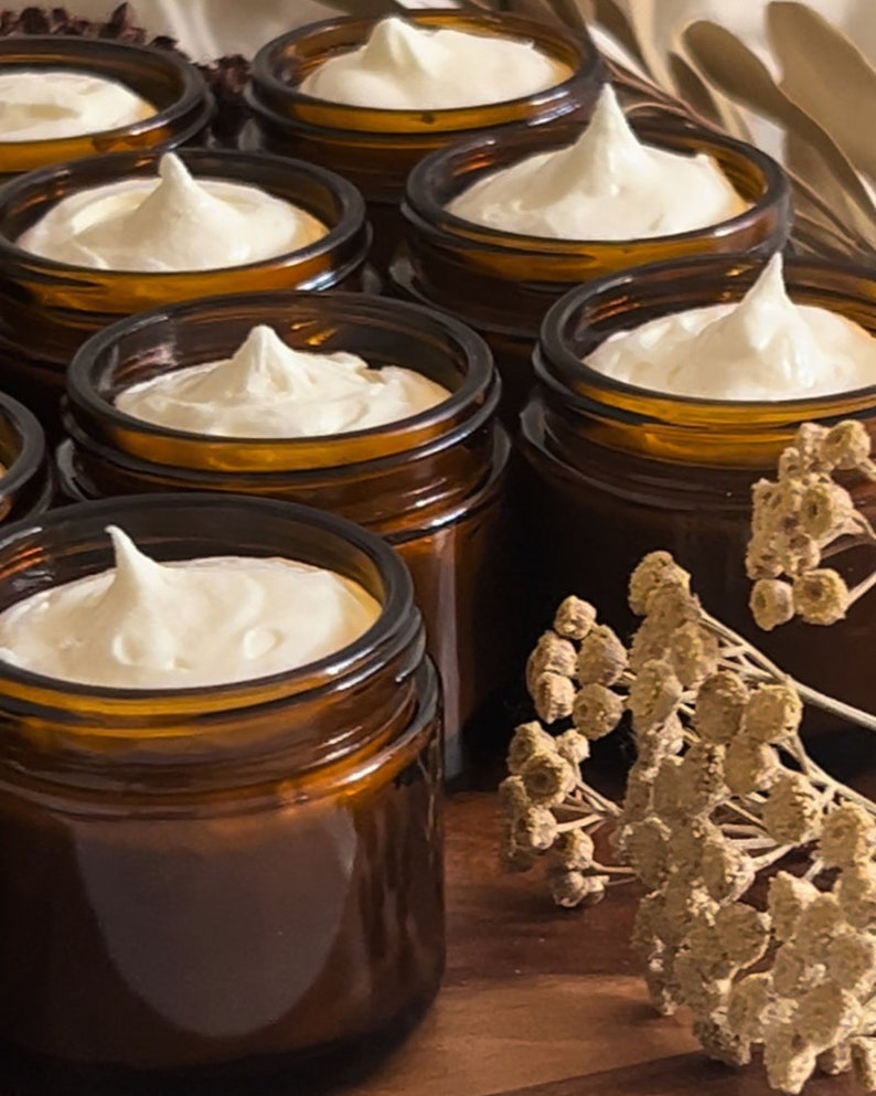May include: Multiple amber glass jars filled with a white cream product. The jars are arranged in a row on a wooden surface with dried flowers in the background.