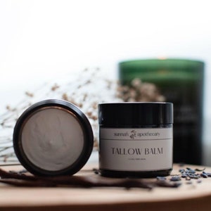 May include: A black jar with a white label that reads "sunnah apothecary TALLOW BALM". The jar is filled with a white balm and is sitting on a wooden surface.