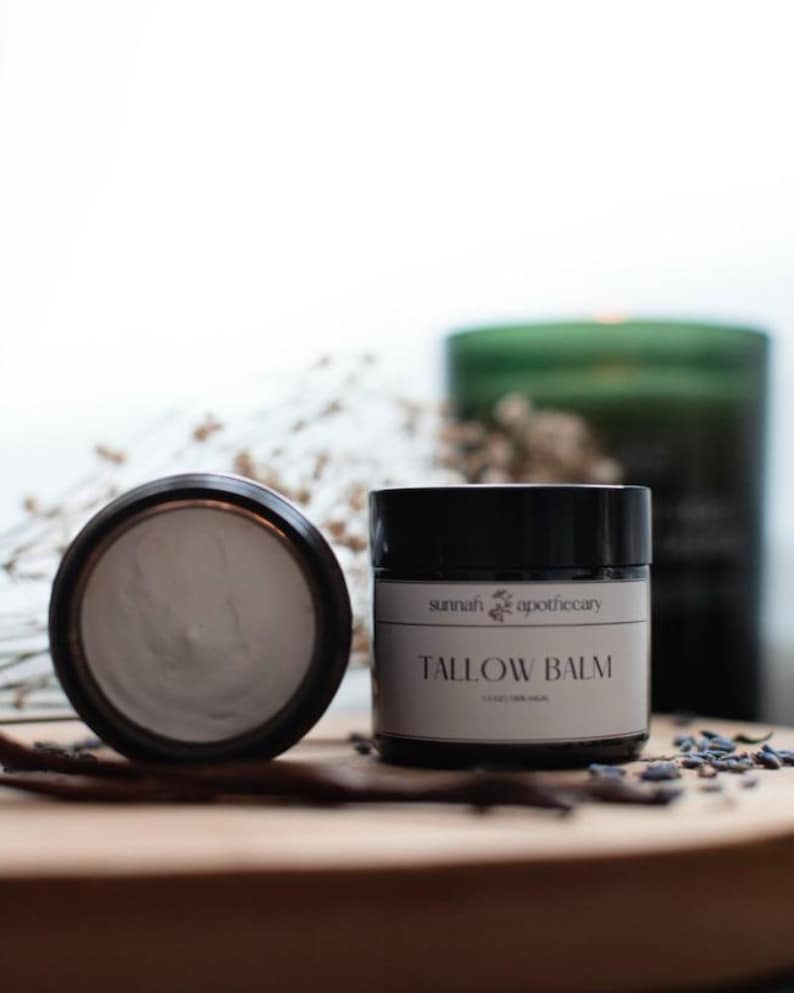 May include: A black jar with a white label that reads "sunnah apothecary TALLOW BALM". The jar is filled with a white balm and is sitting on a wooden surface.