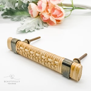 May include: A woven rattan cabinet pull with brass accents. The pull is a light brown colour and has a rectangular shape.