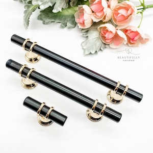 May include: Three black cabinet pulls with gold accents. The pulls have a rectangular shape with rounded edges and two gold rings on the top. The pulls are arranged in a row on a white surface.