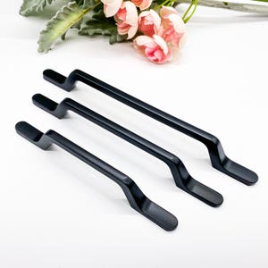 May include: Three black cabinet pulls with a curved design are arranged on a white surface. The pulls are made of metal and have a matte finish. Pink and white flowers are in the background.