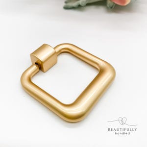 May include: A gold-coloured, square-shaped carabiner with a screw-lock mechanism. The carabiner is made of metal and has a smooth, matte finish. The words "BEAUTIFULLY handled" are printed in black below the carabiner.