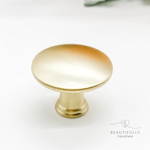 May include: A gold-coloured cabinet knob with a brushed finish. The knob has a rounded, oval shape and a short, cylindrical base. The words "BEAUTIFULLY handled" are printed in black below the knob.
