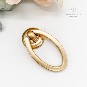 May include: A gold oval-shaped cabinet pull with a ring handle. The pull has a brushed finish.