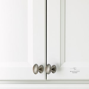Może przedstawiać: Zbliżenie białych drzwi szafki z gałkami ze szczotkowanego niklu. Gałki mają owalny kształt z dekoracyjną krawędzią. Drzwi mają czysty, nowoczesny design. Tekst "BEAUTIFULLY handled" jest wydrukowany na prawych drzwiach.