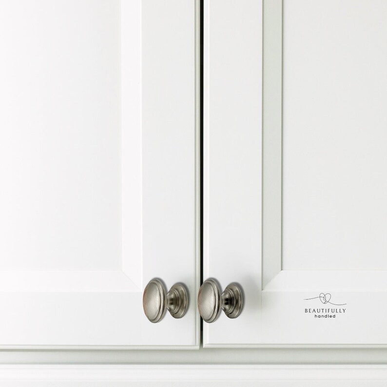 Może przedstawiać: Zbliżenie białych drzwi szafki z gałkami ze szczotkowanego niklu. Gałki mają owalny kształt z dekoracyjną krawędzią. Drzwi mają czysty, nowoczesny design. Tekst "BEAUTIFULLY handled" jest wydrukowany na prawych drzwiach.