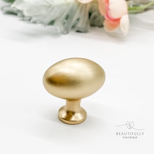 May include: A brushed gold cabinet knob with an oval shape. The knob has a short, cylindrical base and a smooth, rounded top. The hardware is designed for furniture and home decor.