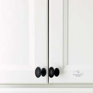 Może przedstawiać: Zbliżenie białych drzwi szafki z matowymi czarnymi owalnymi gałkami szafki. Gałki są wyśrodkowane na drzwiach. Na prawych drzwiach wydrukowano słowa "BEAUTIFULLY handled".