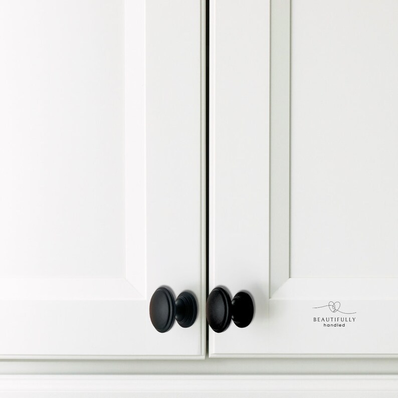 Może przedstawiać: Zbliżenie białych drzwi szafki z matowymi czarnymi owalnymi gałkami szafki. Gałki są wyśrodkowane na drzwiach. Na prawych drzwiach wydrukowano słowa "BEAUTIFULLY handled".