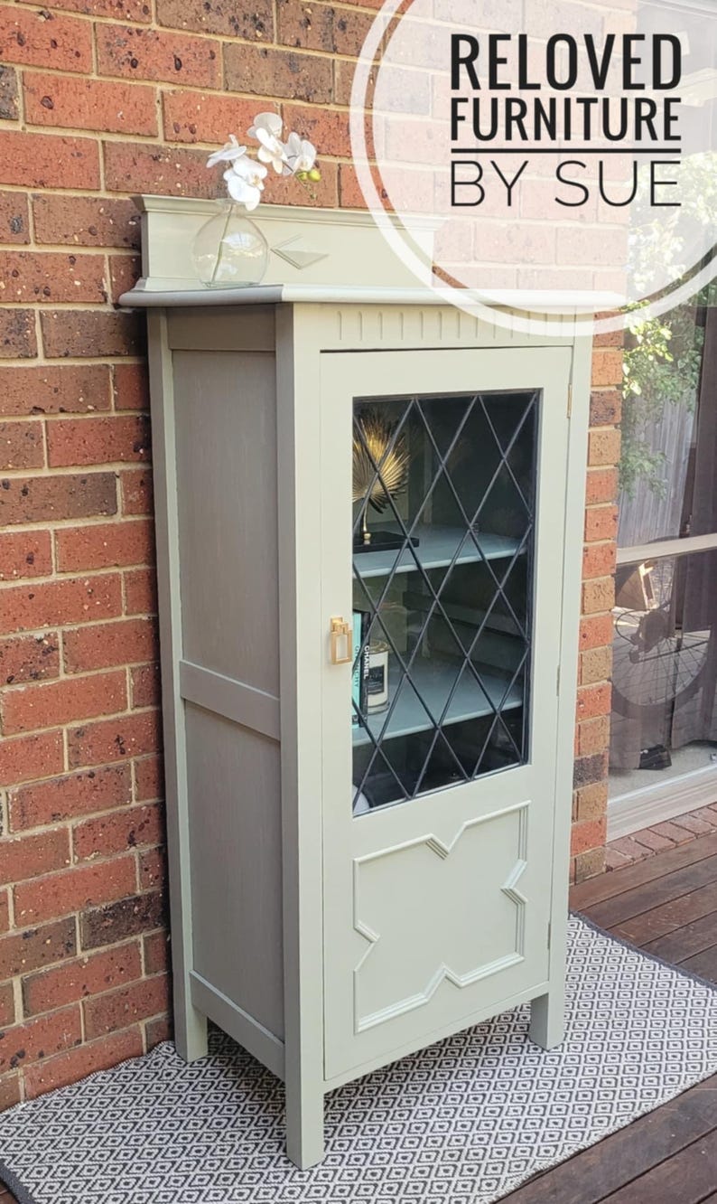 Op de afbeelding: Een lichtgroene kast met een glazen deur en twee planken. De kast heeft een decoratief ontwerp aan de voorkant en staat op een zwart-wit gepatroneerd kleed. De kast is gelabeld met "Reloved Furniture by Sue".