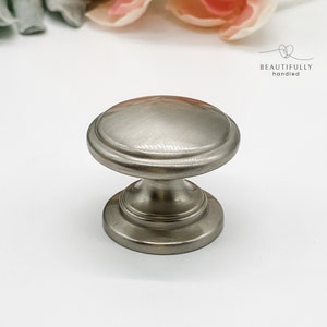 May include: A brushed nickel cabinet knob with a round top and a slightly raised edge. The knob has a simple, elegant design.