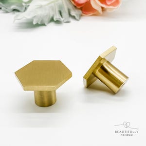 May include: Two brass cabinet knobs with a hexagonal face and cylindrical base. The knobs are a warm gold colour and are set against a white background. The knobs are designed for furniture and home decor.