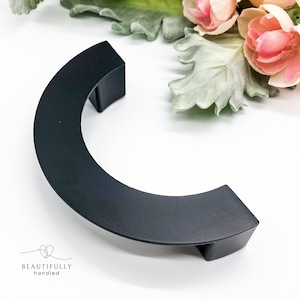 May include: Black C-shaped cabinet hardware with a matte finish. The hardware is made of metal and has a rounded edge.