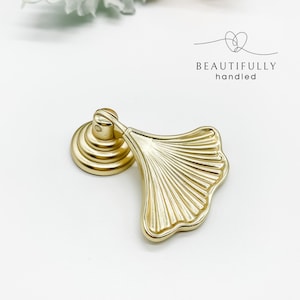 May include: Gold-toned metal cabinet pull with a ginkgo leaf design. The pull has a round base and a leaf-shaped handle.