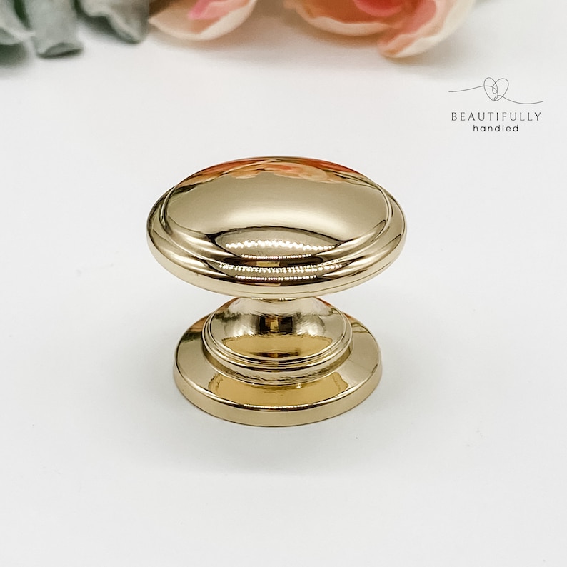 Może przedstawiać: Złoty uchwyt do szafki z zaokrąglonymi g&oacute;rą i podstawą. Gałka ma polerowane wykończenie i dekoracyjny wz&oacute;r. Tekst "BEAUTIFULLY handled" znajduje się w prawym g&oacute;rnym rogu. Odpowiedni do mebli i wystroju domu.