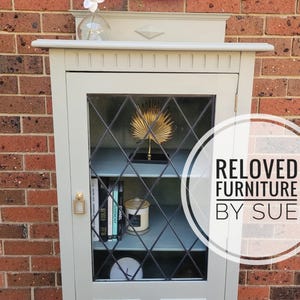 Op de afbeelding: Een lichtgrijze kast met glazen deuren en planken. De kast heeft een ruitvormig patroon op het glas en een gouden palmboomdecoratie op de bovenste plank. De kast is gelabeld met "RELOVED FURNITURE BY SUE".