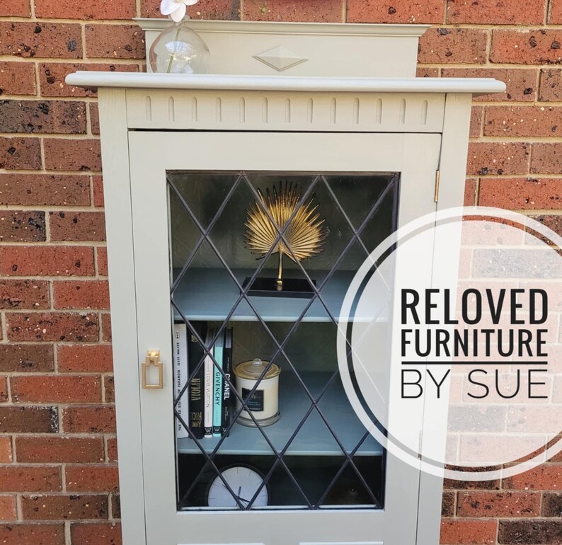 Op de afbeelding: Een lichtgrijze kast met glazen deuren en planken. De kast heeft een ruitvormig patroon op het glas en een gouden palmboomdecoratie op de bovenste plank. De kast is gelabeld met "RELOVED FURNITURE BY SUE".