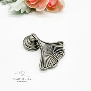 May include: Silver-toned metal cabinet pull with a fan-shaped design. The pull has a round base and a decorative, textured fan-shaped handle.