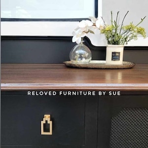 Op de afbeelding: Een zwarte commode met gouden beslag en een houten blad. De commode heeft een zwart-witte gepatroneerde deur aan de rechterkant. De commode is versierd met een vaas met witte bloemen en een kaars op een gouden dienblad. De tekst "RELOVED FURNITURE BY SUE" is gedrukt op de voorkant van de commode.