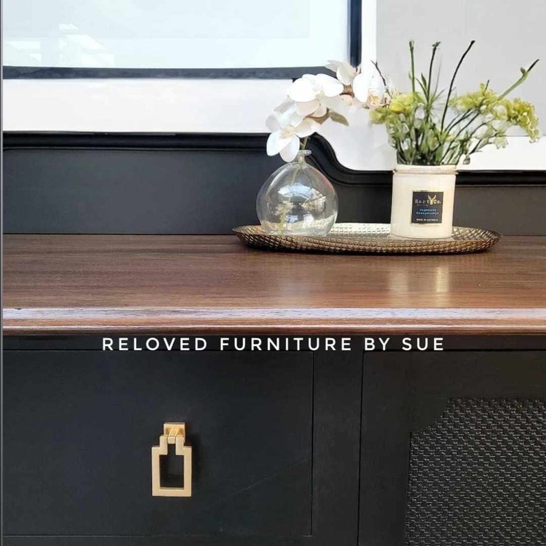 Op de afbeelding: Een zwarte commode met gouden beslag en een houten blad. De commode heeft een zwart-witte gepatroneerde deur aan de rechterkant. De commode is versierd met een vaas met witte bloemen en een kaars op een gouden dienblad. De tekst "RELOVED FURNITURE BY SUE" is gedrukt op de voorkant van de commode.