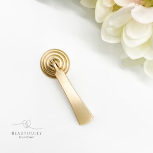 May include: A gold-toned cabinet pull with a rounded, ridged top and a long, straight handle. The handle has a slightly curved end.