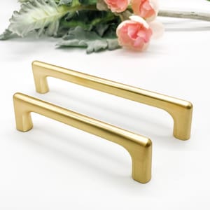 May include: Two gold cabinet pulls with a sleek, minimalist design. The pulls are rectangular with rounded edges and a brushed finish. The hardware is shown against a white background with floral accents.