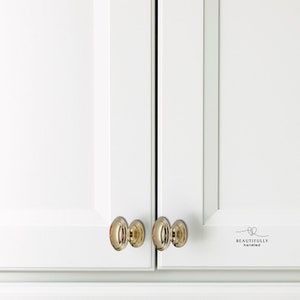 Może przedstawiać: Zbliżenie białych drzwi szafki z polerowanymi, złotymi, owalnymi gałkami. Gałki są wyśrodkowane na drzwiach, kt&oacute;re są lekko uchylone. Tekst "BEAUTIFULLY handled" jest wydrukowany na prawych drzwiach.