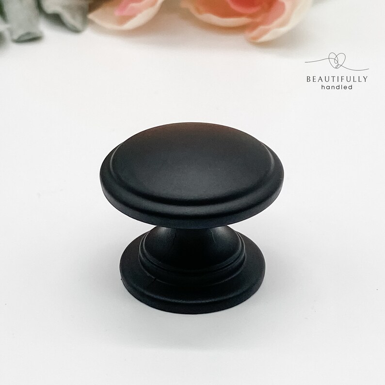 Może przedstawiać: Matowy czarny uchwyt do szafki z zaokrąglonym blatem i dekoracyjną podstawą. Uchwyt jest wyśrodkowany na białej powierzchni. Tekst "BEAUTIFULLY handled" znajduje się w prawym g&oacute;rnym rogu.