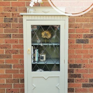 Op de afbeelding: Een saliegroene kast met glazen deuren en een ruitvormig patroon. De kast heeft een plank binnenin en is versierd met een gouden waaier en boeken. De kast staat op een zwart-wit gepatroneerd tapijt.