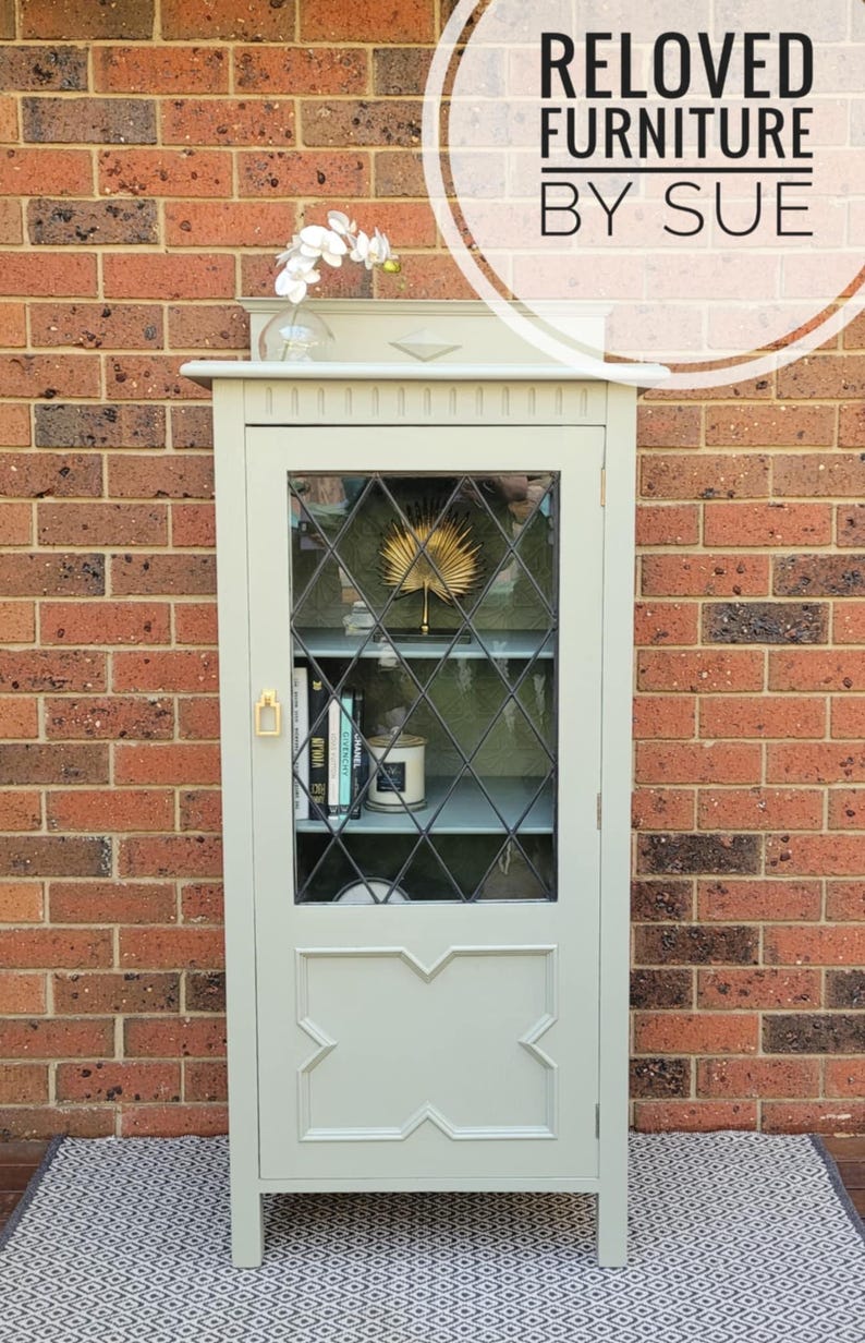 Op de afbeelding: Een saliegroene kast met glazen deuren en een ruitvormig patroon. De kast heeft een plank binnenin en is versierd met een gouden waaier en boeken. De kast staat op een zwart-wit gepatroneerd tapijt.