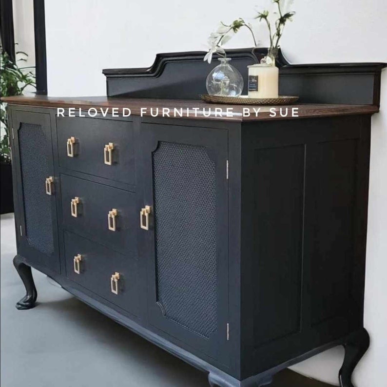 Op de afbeelding: Een antiek dressoir, zwart geverfd, met drie lades en twee deuren. De deuren hebben een zwarte gaas inzet. Het dressoir heeft goudkleurige beslag en staat op vier poten. De bovenkant van het dressoir is van donker hout.