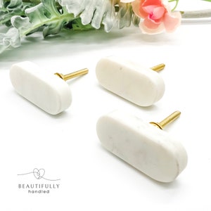 May include: Three white marble cabinet knobs with an elongated oval shape and a smooth finish. Each knob features a gold-coloured metal threaded bolt and a small washer. The text "BEAUTIFULLY handled" is printed in black in the bottom left corner.