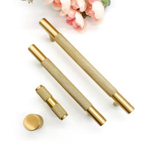 May include: A set of gold-coloured cabinet hardware, comprising two long bar pulls, a shorter pull, and a round knob. The hardware has a textured, diamond-patterned grip and smooth, cylindrical ends. Pink flowers are in the background.