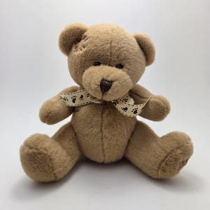May include: A plush brown teddy bear with a light brown bow featuring paw prints. The bear has a stitched patch on its head. The bear is sitting with its arms and legs spread out.