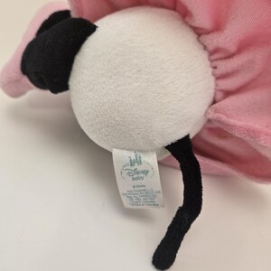 Puede incluir: Un juguete de peluche de flamenco blanco con un tut&uacute; rosa y ojos negros. El juguete tiene una etiqueta que dice "Disney Baby" e incluye el logotipo de Disney.