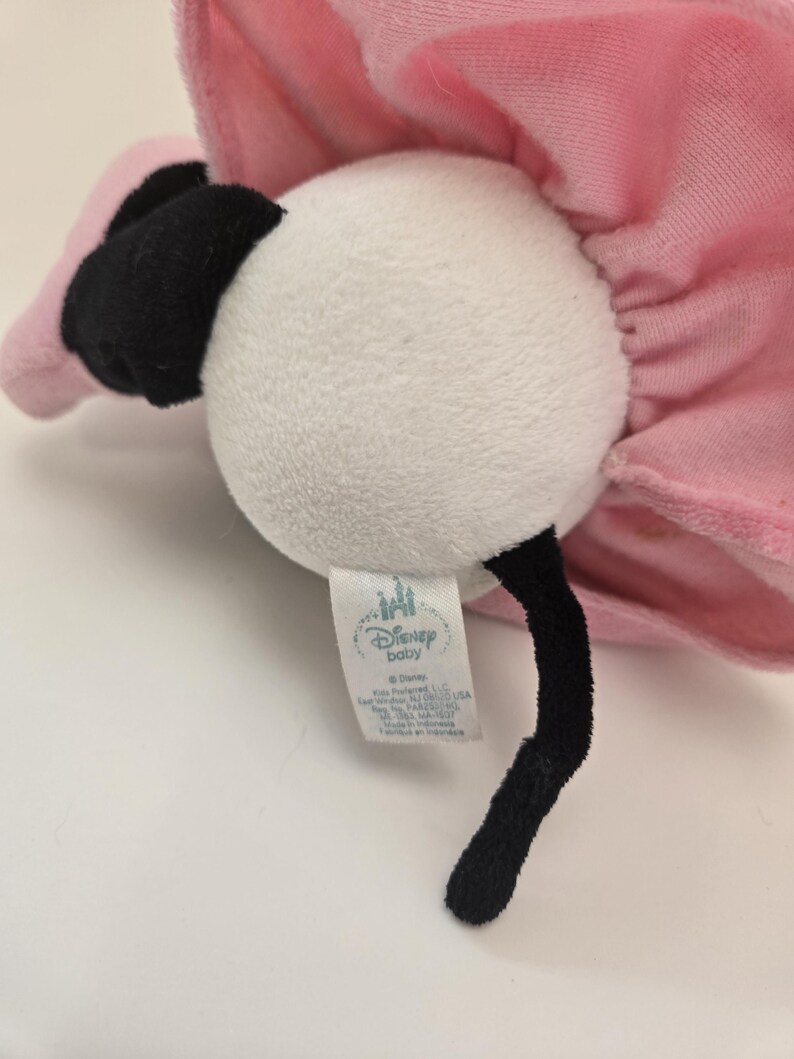 Puede incluir: Un juguete de peluche de flamenco blanco con un tut&uacute; rosa y ojos negros. El juguete tiene una etiqueta que dice "Disney Baby" e incluye el logotipo de Disney.