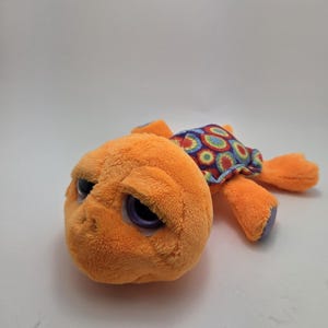May include: A plush orange turtle toy with large, expressive eyes. The turtle's shell is patterned with colorful circles. The toy is lying on a white surface.
