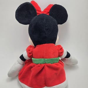 May include: A red and white plush Minnie Mouse toy wearing a green belt and a red bow. The toy is sitting on a white background.