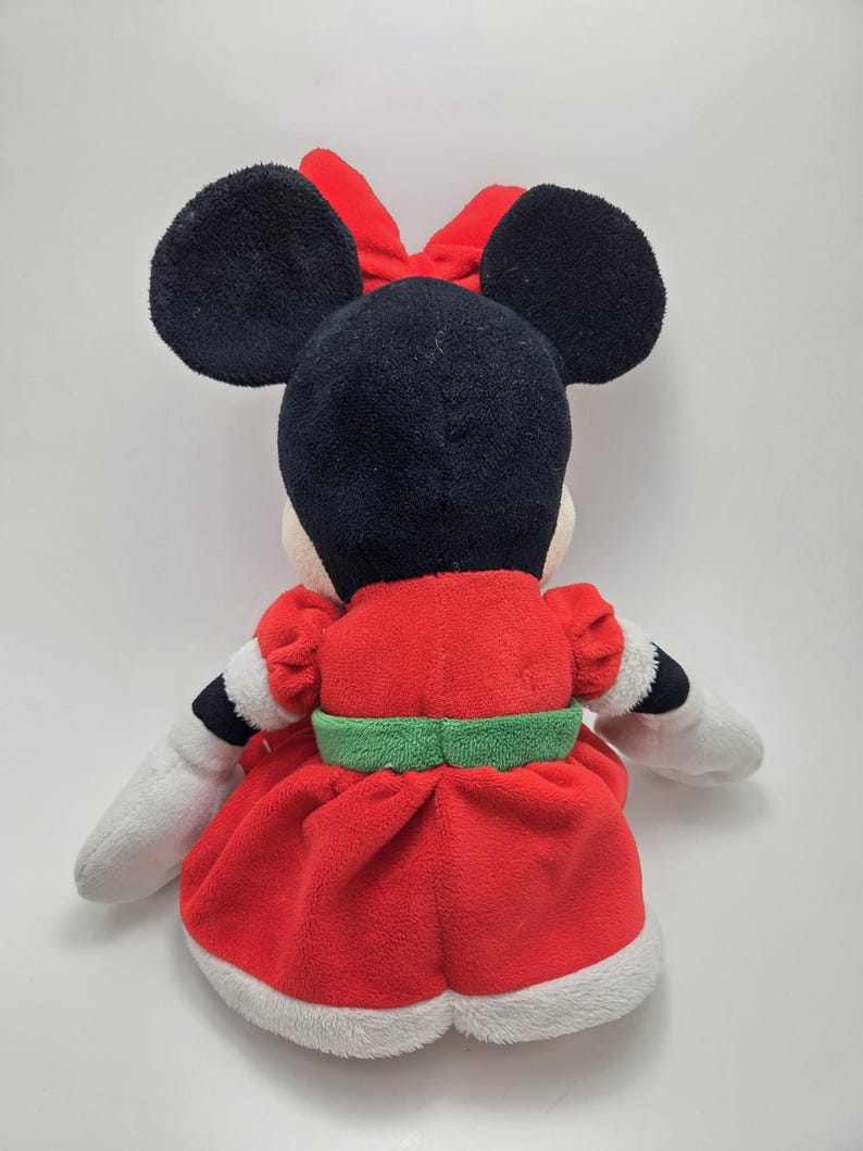 May include: A red and white plush Minnie Mouse toy wearing a green belt and a red bow. The toy is sitting on a white background.