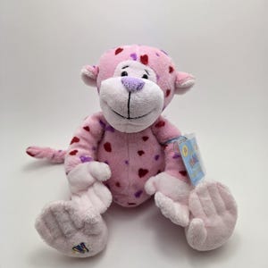 May include: A pink plush monkey toy, decorated with red and purple heart shapes. The monkey features a white face, purple nose, and a tag. The toy is designed with a smiling expression and soft, plush material.