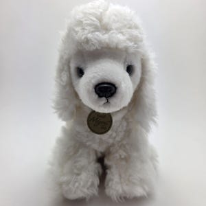 May include: A white plush poodle toy with a fluffy, curly coat. The toy has a black nose, dark eyes, and a small, round tag. The poodle is sitting upright, showcasing its soft, textured fur.