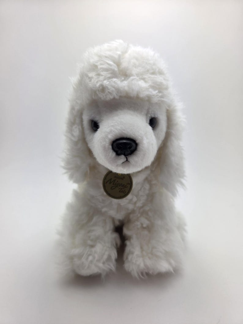 May include: A white plush poodle toy with a fluffy, curly coat. The toy has a black nose, dark eyes, and a small, round tag. The poodle is sitting upright, showcasing its soft, textured fur.