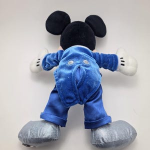 May include: A plush Mickey Mouse toy, viewed from the back, with black ears and head, white gloves, and silver shoes. The toy is wearing a blue velvet jacket and pants. The toy's back is facing the viewer.