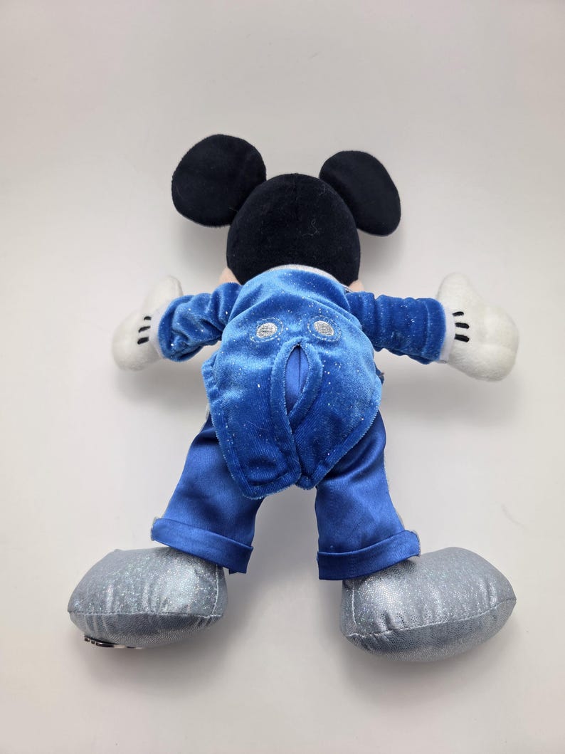 May include: A plush Mickey Mouse toy, viewed from the back, with black ears and head, white gloves, and silver shoes. The toy is wearing a blue velvet jacket and pants. The toy's back is facing the viewer.