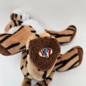 Puede incluir: Un tigre de peluche rayado marr&oacute;n y negro con vientre blanco. El tigre tiene una pata marr&oacute;n con un logotipo "Ty" colorido en ella.